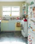 Retro kitchen with pastel cabinets, patterned wallpaper, fridge with magnets, and a red kettle on the stove.