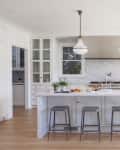 Spacious kitchen with marble island, four stools, pendant lights, and glass cabinets. Dining table with chairs by large windows.