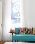 Turquoise sofa with colorful patchwork quilt, side table with stacked books, and sheer curtains framing a window view.