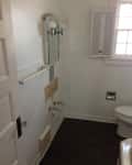 Bathroom with missing sink, wall-mounted mirror, light fixtures, and a toilet next to a window with shutters.