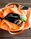 Orange gym bag with a towel, water bottle, and yellow dumbbells on a wooden floor.