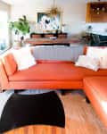 Orange sectional sofa with patterned throw, fluffy pillows, wooden coffee table, and dining area with abstract art and chandelier.