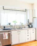 Bright kitchen with white cabinets, marble countertops, open shelves, and a window with potted plants.
