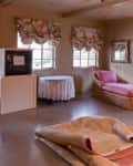 Room with wicker chaise lounges, floral curtains, a TV on a stand, and a rolled-up carpet on the floor.