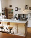 Vintage kitchen with white cabinets, marble island, wooden stools, and retro decor, including a wall clock and hanging lights.