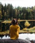 Person in a yellow shawl and hat sitting on a rock, overlooking a forested lake.