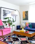 Living room with a navy sofa, pink armchair, colorful rug, and plants, featuring framed art and a wooden coffee table.