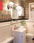 Small bathroom with floral wallpaper, white tiles, vanity, mirror, and wall sconces.