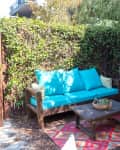 Outdoor patio with turquoise sofa, pink chair, wooden table, and colorful rug surrounded by greenery and bamboo fencing.