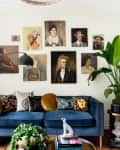 Blue velvet sofa with colorful pillows, surrounded by plants, under a gallery wall of diverse portraits.