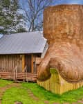 Wooden cabin with a large boot-shaped structure attached, surrounded by trees and grass.