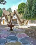 Whimsical cottage with curved roofs, stone facade, colorful stone patio, and central fountain surrounded by lush greenery.