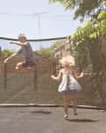 Two children jumping on a backyard trampoline surrounded by trees and a wooden fence.