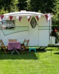 Vintage caravan with bunting and lanterns, surrounded by greenery, with a table and chairs on the grass.