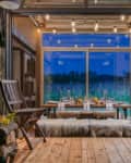 Rustic dining area with wooden chairs, a table set for dinner, and string lights, overlooking a field at dusk.