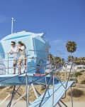 Two people standing on a blue lifeguard tower overlooking a sandy beach with palm trees.