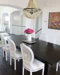 Dining room with dark wood table, white chairs, pink flowers in vase, beaded chandelier, and colorful wall art.