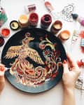 Hands painting a phoenix on a black plate surrounded by colorful paint jars and art supplies.