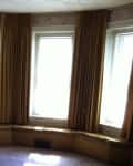 Bay window with brown curtains, floral wallpaper, and a floor lamp in a dimly lit room.