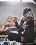 Woman in patterned headscarf holding a steaming glass of tea, sitting on a floor with patterned cushions and a tray with a teapot.