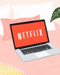 Laptop displaying Netflix on a pink bed with decorative pillows, a plant, and a side table with a bottle and cream tube.