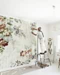Man applying floral wallpaper in a bright room, standing on a ladder.