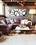 Cozy living room with brown leather sectional, abstract art, floor lamp, bean bag, and cowhide rug under string lights.