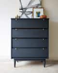 Dark blue dresser with four drawers, gold handles, abstract art, candles, and a small plant on top.