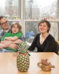 Family of four sitting at a table with a pineapple, teapot, and coasters, smiling in front of large windows.