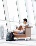 Man sitting in a wicker chair at an airport terminal, wearing headphones, with a backpack on the floor.
