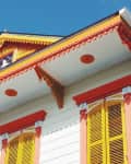 Colorful Caribbean-style house facade with yellow shutters, red and orange trim, and decorative eaves against a blue sky.