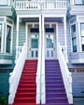 Two staircases, one red and one purple, lead to a light blue house with white trim and a small balcony.