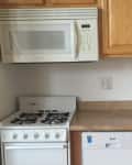 Compact kitchen with white microwave, stove, dishwasher, and small sink under wooden cabinets.