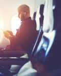 Man sitting in airplane seat, using smartphone with earphones, sunlight streaming through window.