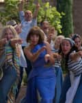 Group of people dancing outdoors, wearing colorful outfits, pointing forward, with stone buildings and greenery in the background.