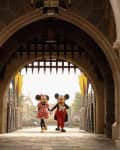 Mickey and Minnie Mouse walking hand in hand under a stone archway at a theme park entrance.
