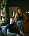 A man with tattoos sits in a chair, and a woman stands beside him, both smiling in a room with plants and wooden furniture.