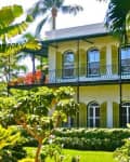 Two-story yellow house with green shutters, surrounded by lush tropical plants and palm trees.
