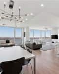 Open-concept living area with modern chandelier, marble kitchen island, and large windows offering city views.