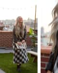 Woman with long hair in plaid dress and leather jacket on rooftop, holding a clutch, with cityscape in background.