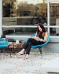 Woman reading on a blue chair in a patio with a stone table, colorful cushions, and large windows.