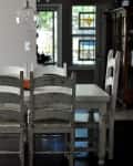 Rustic dining room with white table, six distressed wooden chairs, pendant light, and stained glass window.
