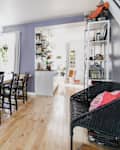 Dining room with wooden table, black chairs, purple walls, and framed art, leading to a bright kitchen with plants.
