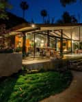 Modern glass-walled house with outdoor patio, lush greenery, and warm interior lighting at night.