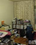 Cluttered bedroom with floral curtains, messy bed, bookshelves, and scattered clothes.