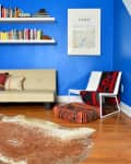 Blue-walled room with a beige sofa, colorful bookshelves, Paris poster, patterned chair, and cowhide rug on wooden floor.