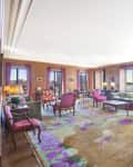 Spacious living room with colorful furniture, large windows, vibrant rug, and views of the city skyline and river.