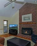 Living room with brown leather sofa, brick fireplace, TV, and large windows overlooking a garden.