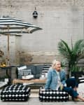 Outdoor patio with black and white furniture, striped umbrella, plants, and a woman sitting on a patterned ottoman.