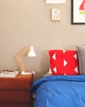 Bed with blue duvet, red pillow with white triangles, wooden lamp on nightstand, and framed art on beige wall.